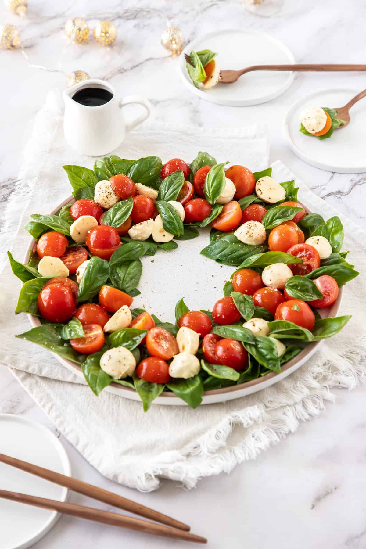 Caprese salad in a wreath circle, on a white plate, sitting on a white cloth, with a jug of balsamic glaze in the background.