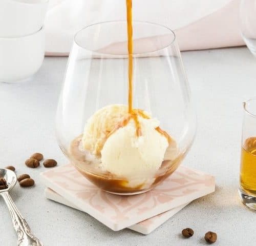 glass with ice cream and coffee being poured over, sitting on a napkin, with a shot of liqueur, a spoon and some coffee beans around the edge.