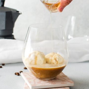 glass with ice cream and coffee and liqueur being poured over, with some coffee beans and a spoon on edge and a coffee pot in the background.
