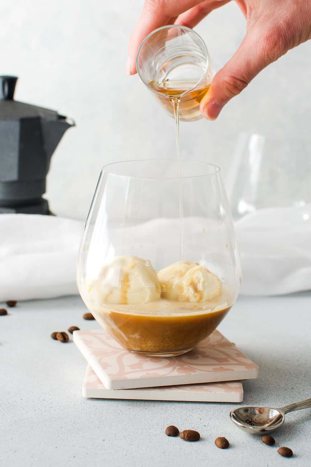 glass with ice cream and coffee and liqueur being poured over, with some coffee beans and a spoon on edge and a coffee pot in the background.