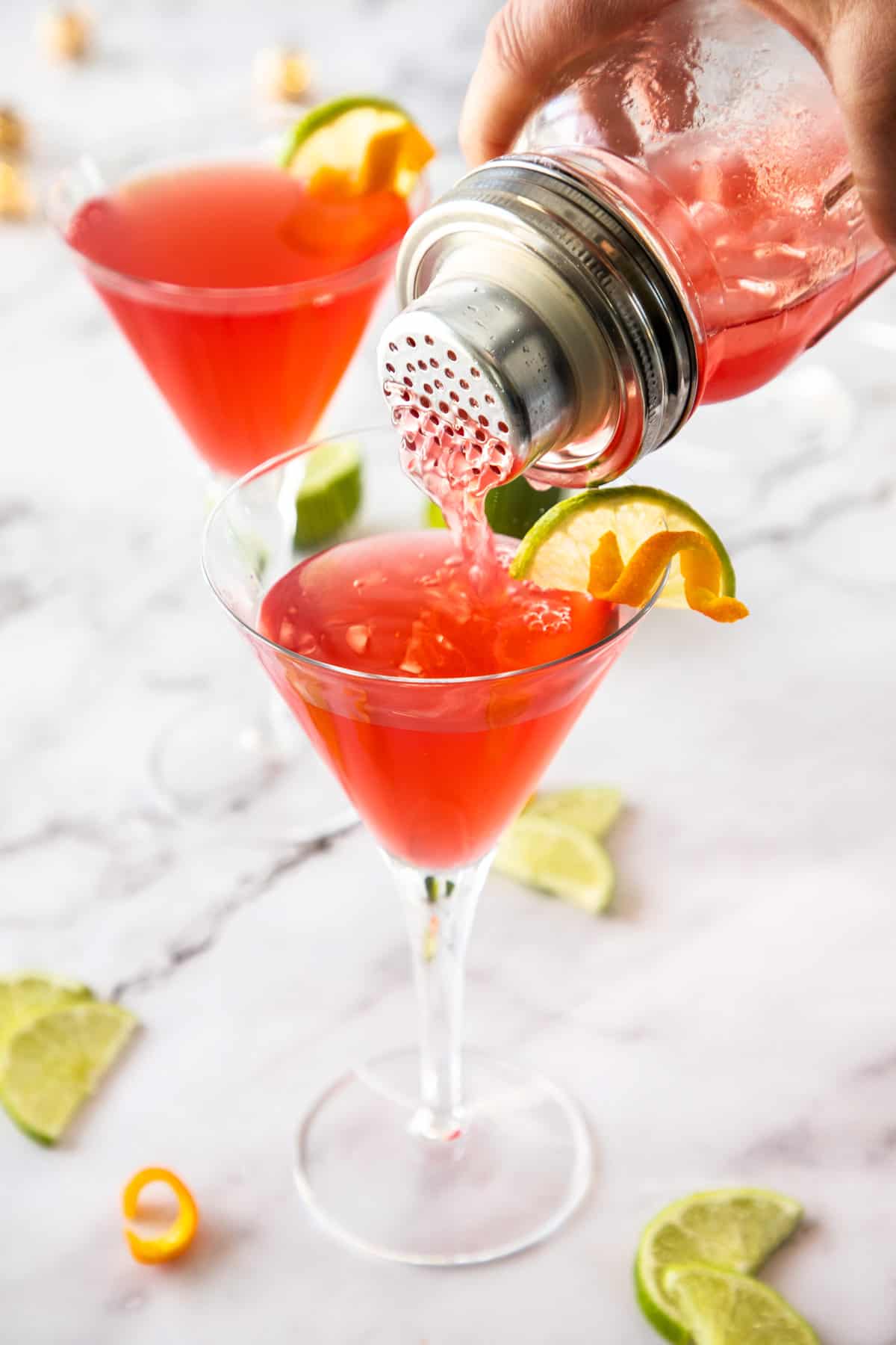 A cocktail shaker pouring a red cocktail into a martini glass.