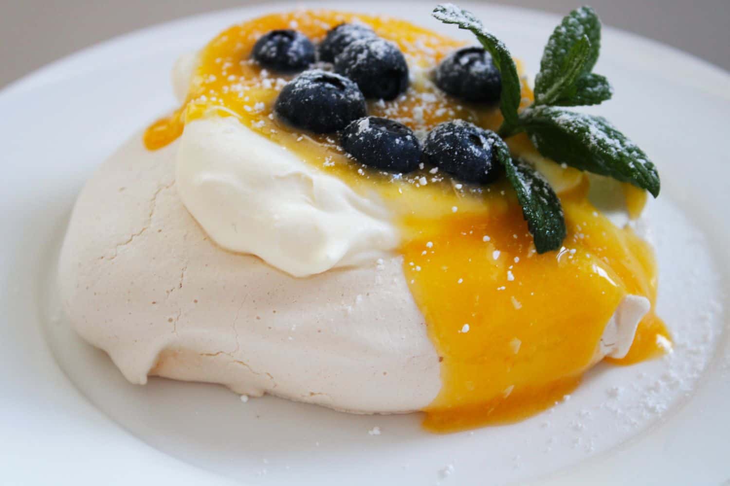 One Meringue on a white plate, topped with cream, lemon curd and blueberries. Garnished with a sprig of mint. 