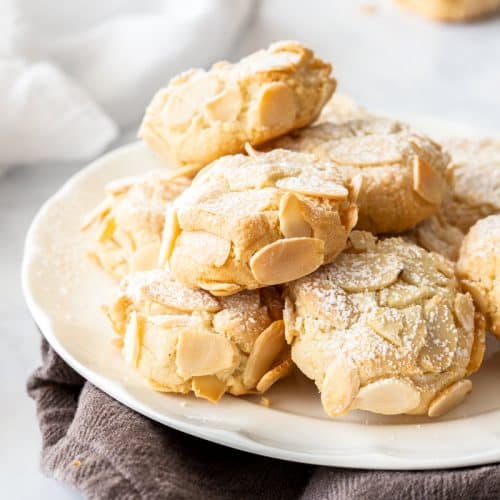 plate of almond cookies on a brown cloth. a cookie and white cloth sit in the background.