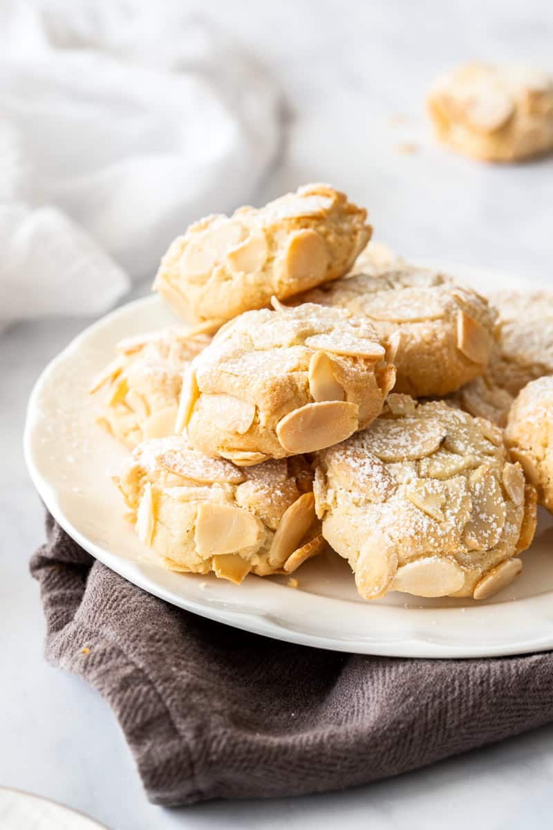 plate of almond cookies on a brown cloth. a cookie and white cloth sit in the background.