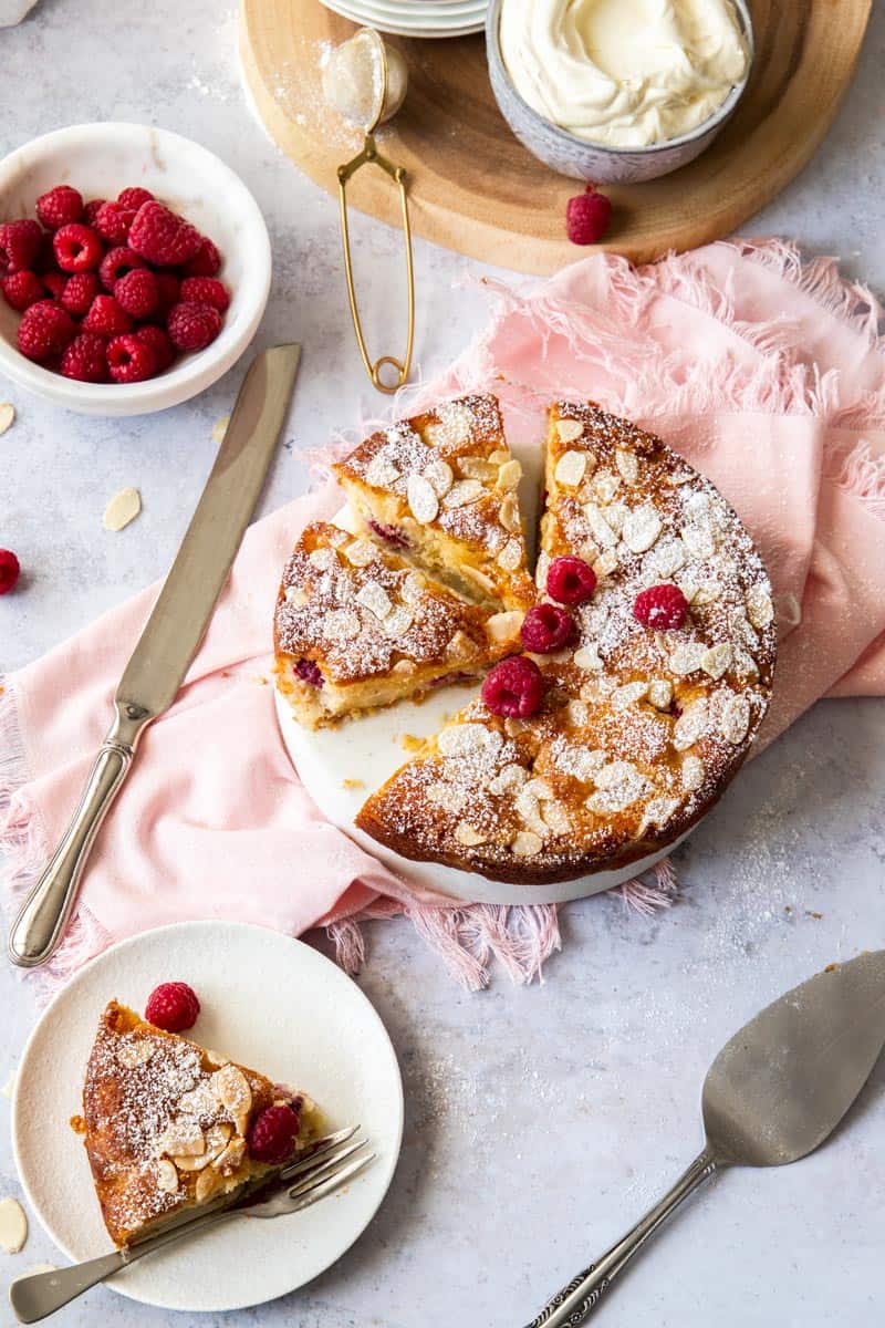 pear cake in centre, with a slice on the left. serving utensils and a bowl of raspberries and cream sit at the top of the photo. cake sitting on pink shawl.