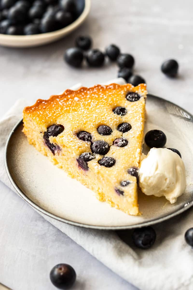 Slice of Gluten-Free Blueberry Cake on grey plate with a dollop of cream.