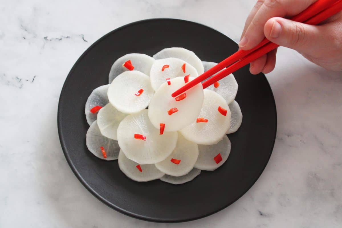 slices of daikon on a black plate, garnished with a little finely chopped red chilli. A piece is being picked up with some red chopsticks.