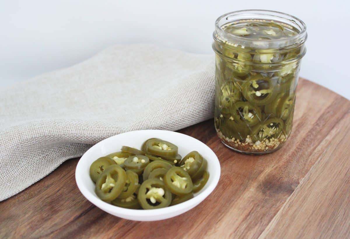 small white round dish of jalapeno and a jar of jalapeno, on a wooden board, with a pale brown napkin.