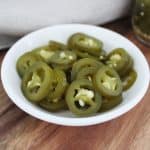 small white round dish of jalapeno on a wooden board, with a pale brown napkin.