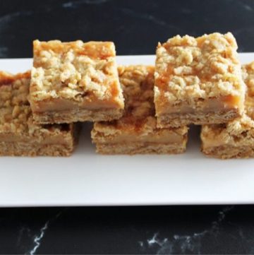 five caramel crumb bars on a white plate, sitting on a black benchtop.