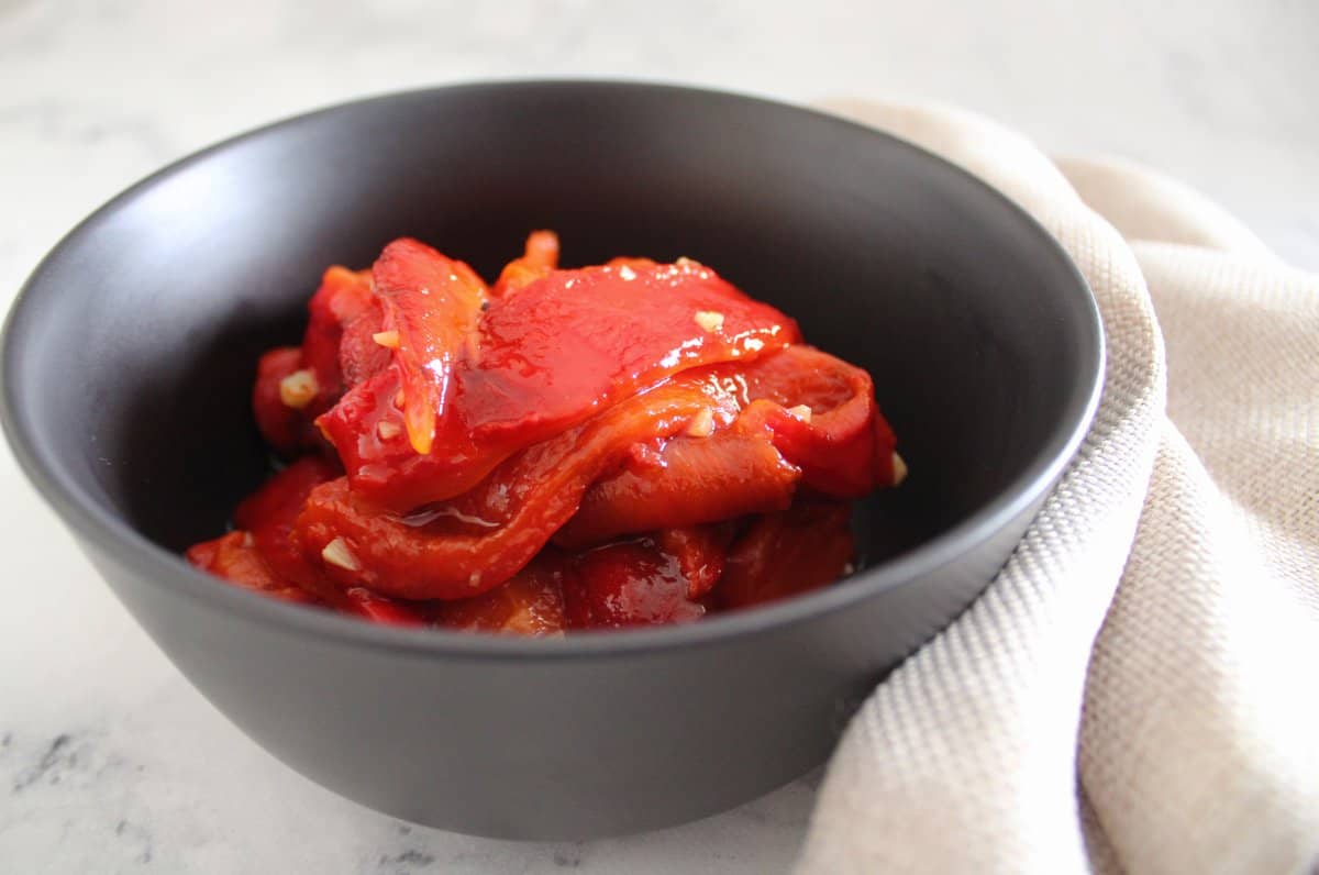 black bowl with roasted capsicums, with a beige napkin. Sitting on a marble benchtop.