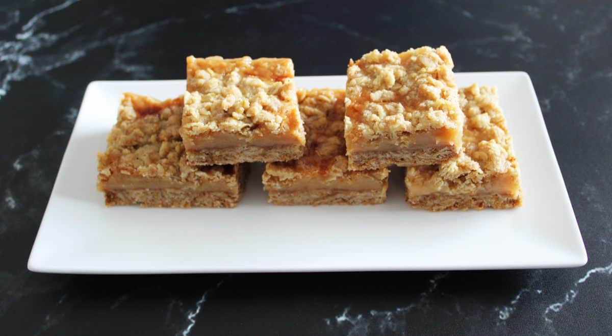 Five caramel crumb bars on a white plate on a black benchtop.