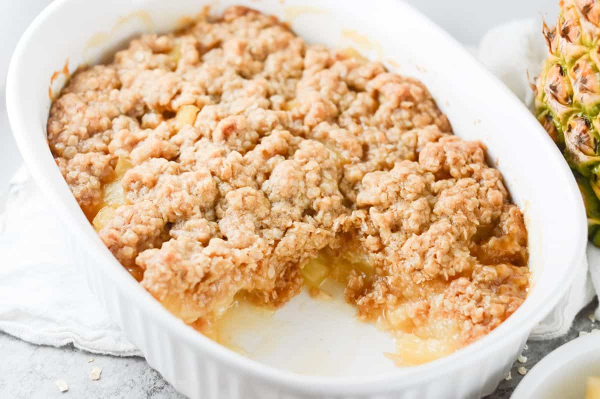 up close shot of baked pineapple crisp, with a serve missing, in a white dish. 