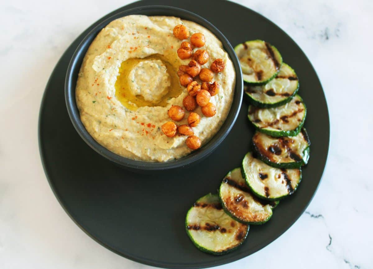 On a black plate, a bowl of hummus in a black bowl. Garnished with a drizzle of oil and crispy chickpeas. Surrounded by some grilled zucchini slices on the plate.