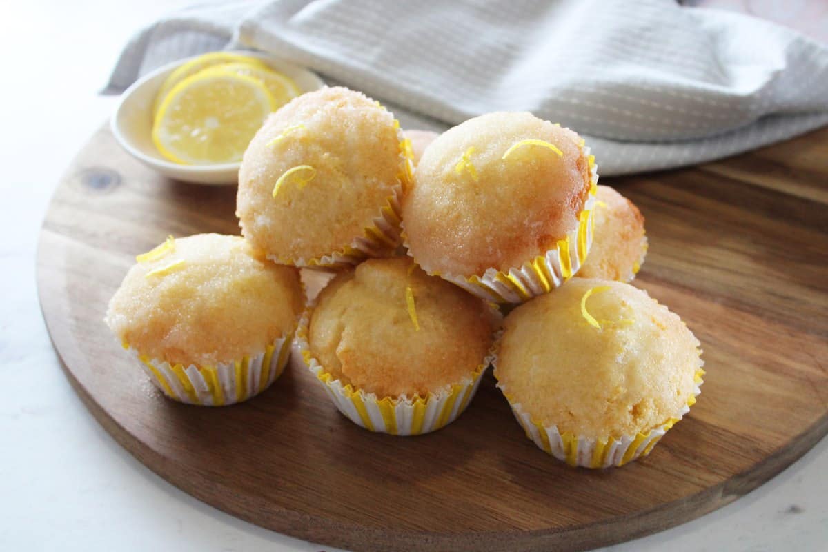 lemon drizzle cupcakes piled up on a round wooden board. a small round dish of lemon slices and a grey tea towel in the background.