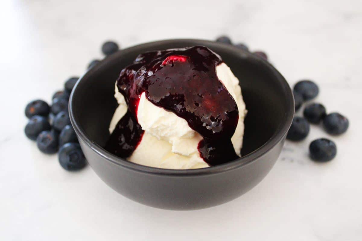 small round black bowl with yoghurt, topped with blueberry sauce, fresh blueberries scattered around the bowl.
