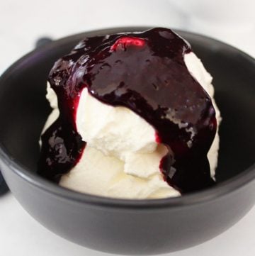 small round black bowl with yoghurt, topped with blueberry sauce, fresh blueberries scattered around the bowl.