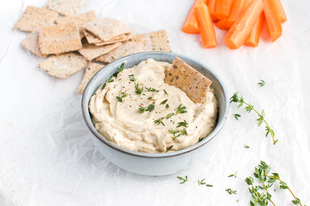 bowl of onion dip, on a white cloth. some crackers and carrots sit behind the bowl, and some thyme is sprinkled around the edge.