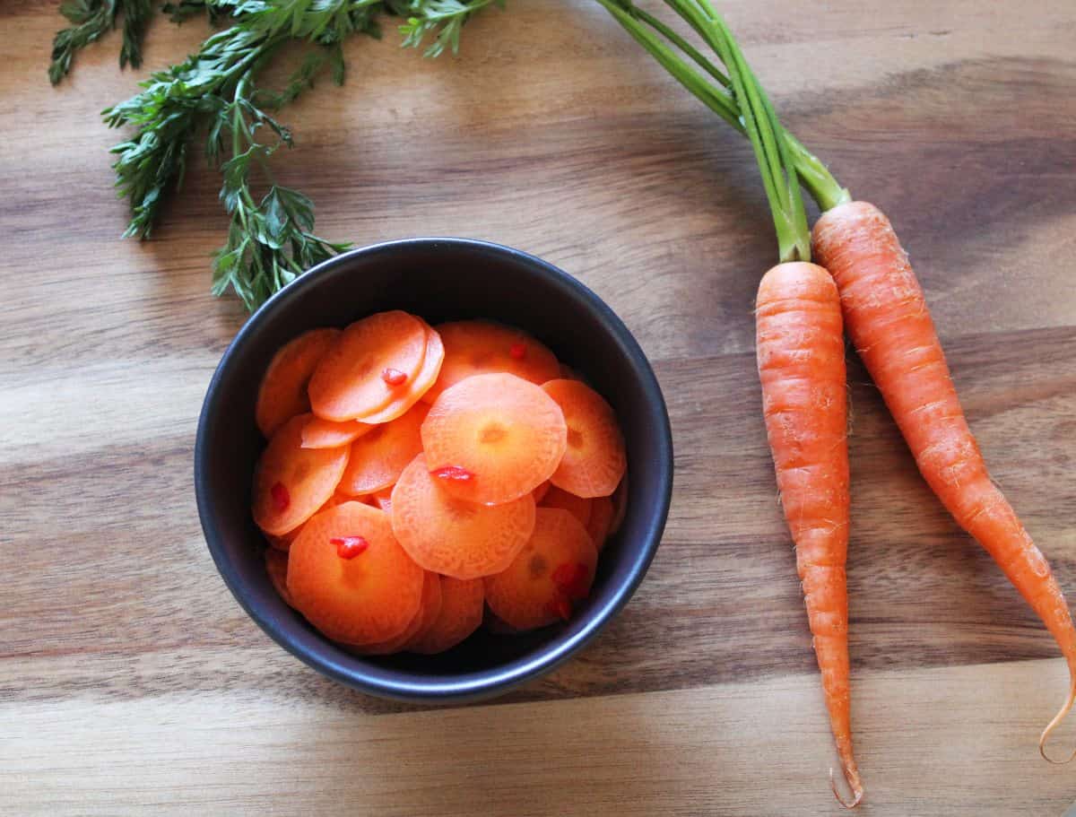 on a round wooden board, a round black bowl of pickled carrots, with two carrots on the right.