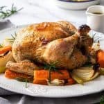 Roast Chicken on a white platter, surrounded by carrot, celery and onion. A sprig of rosemary and white jug of gravy are in the background.