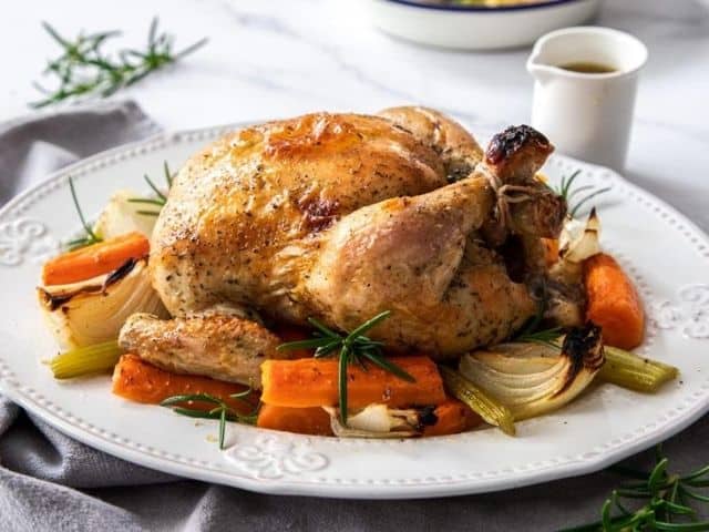 Roast Chicken on a white platter, surrounded by carrot, celery and onion. A sprig of rosemary and white jug of gravy are in the background.