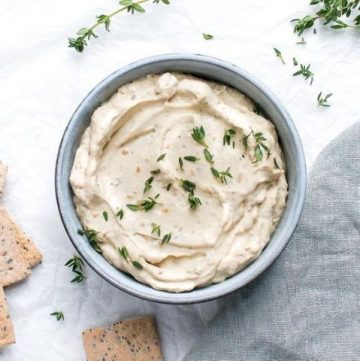 onion dip, on a white cloth background, with some crackers to the left of the bowl, and some sprigs of thyme to the right, a grey cloth in the right corner.