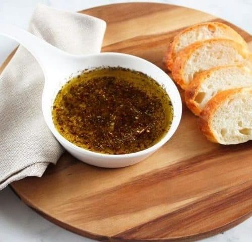 On a round wooden board, a beige napkin, round white dish of dipping oil and four slices of bread.
