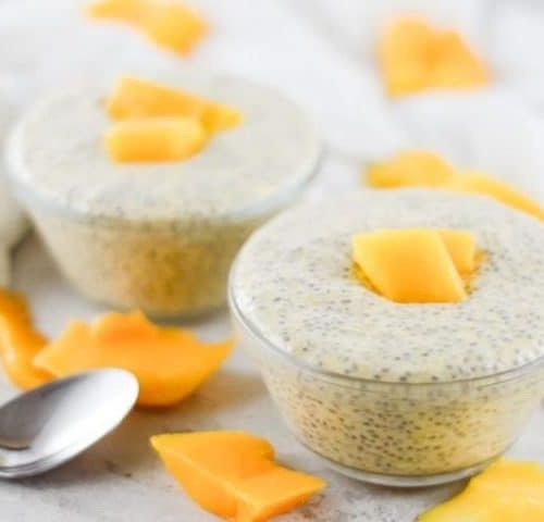 On a white bench, two bowls of chia pudding, garnished with mango sit. Mango pieces scattered around the bowls, and there is a silver spoon on the left. A white cloth in the background.