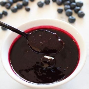 Small white dish of blueberry sauce, with a black spoon dipping into sauce. Some fresh blueberries scattered in the background.