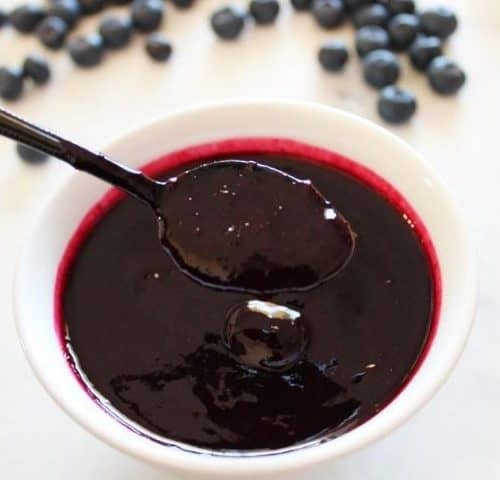 Small white dish of blueberry sauce, with a black spoon dipping into sauce. Some fresh blueberries scattered in the background.