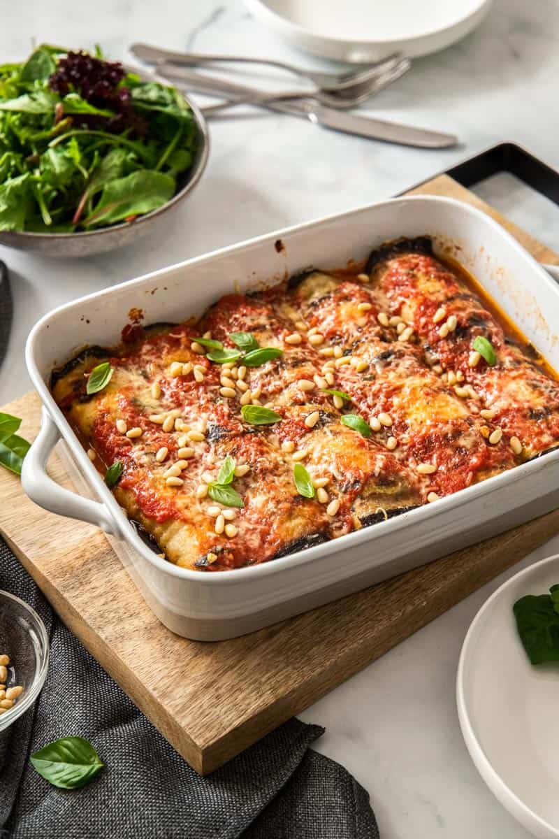 baking dish full of involtini, sitting on a wooden board, with some basil leaves around the edge. a plate and some cutlery and a dish of salad greens sit in the background.