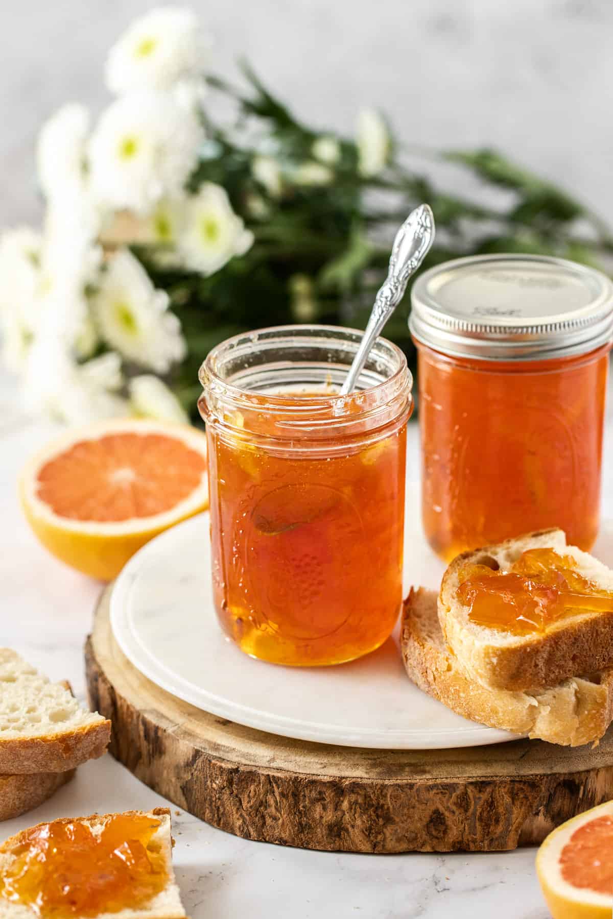 two jars of marmalade on a round board, one with a spoon in it, and some bread and grapefruit surrounding the board.