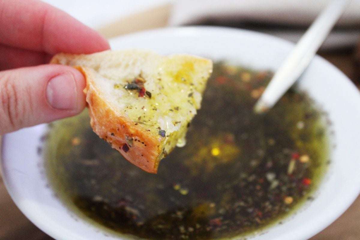 Hand holding a small piece of bread which has been dipping into a round white bowl of dipping oil. Silver spoon in bowl.