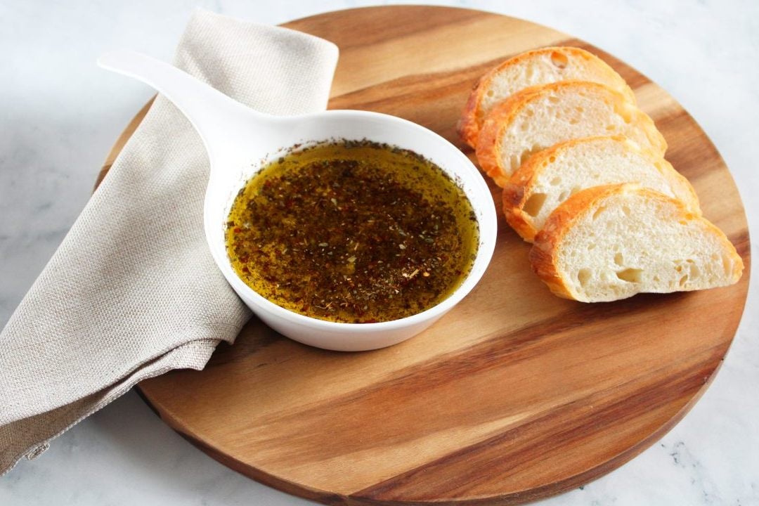 On a round wooden board, a beige napkin, round white dish of dipping oil and four slices of bread.