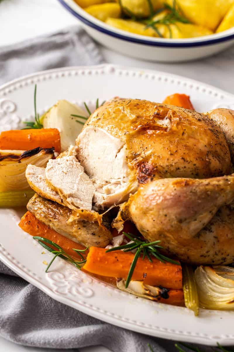 Up close shot of Roast Chicken on a white platter, surrounded by carrot, celery and onion. A piece of the meat has been carved.