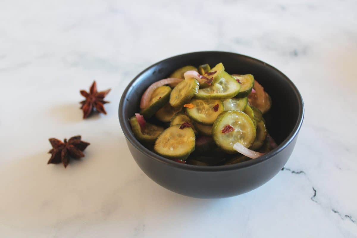Round black bowl of spicy pickles, on a white marble background. Two star anise to the left of the bowl.