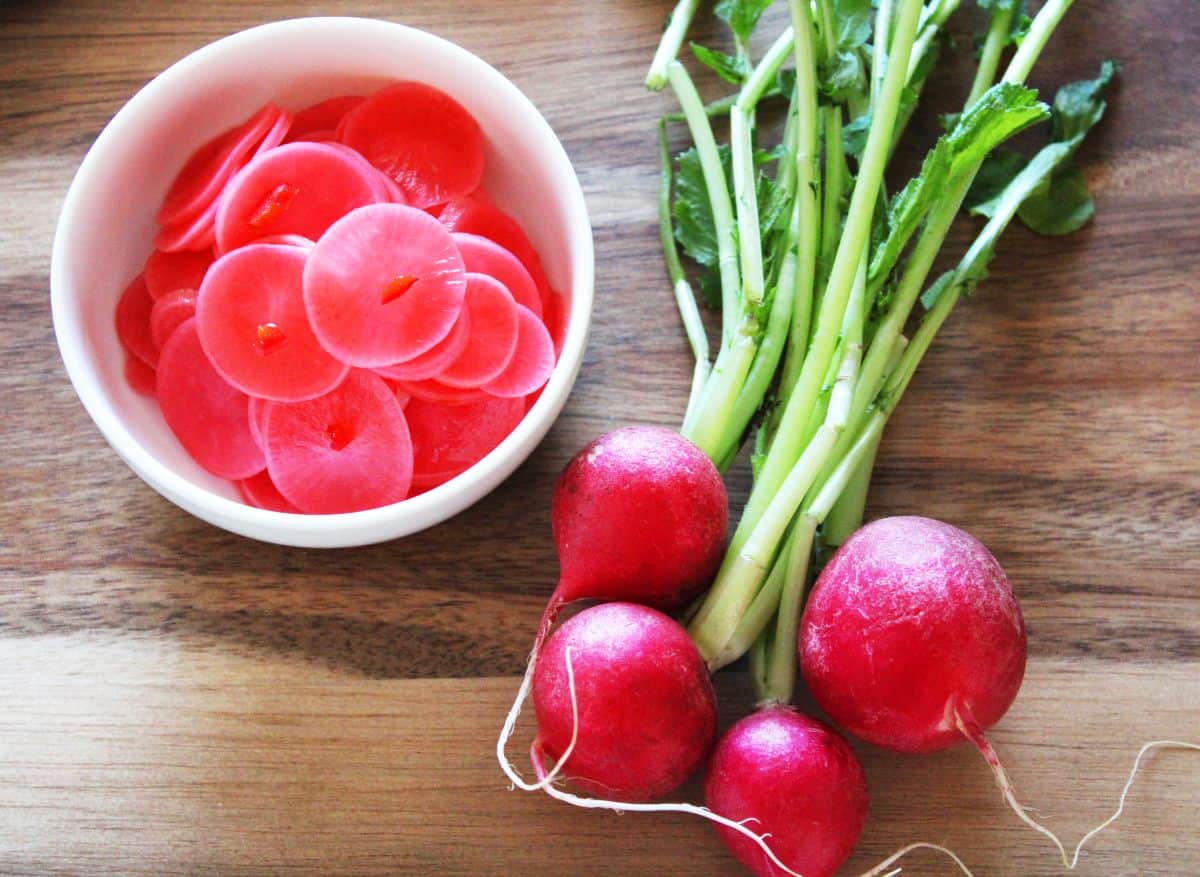 Round white bowl of pickled radishes and a bunch of radishes to the right of bowl, on a wooden board.