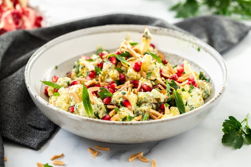 round bowl of salad, sitting on a blue cloth. in the background, some pomegranate and parsley. photo taken on a white marble bench top.