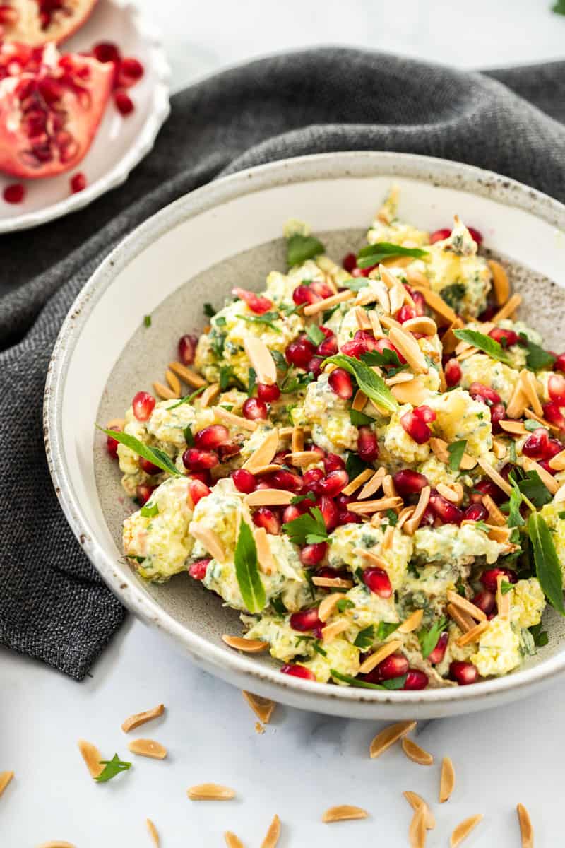 up close shot of bowl of cauliflower salad, sitting on a blue cloth. some slivered almonds are sprinkled at the front, and half a pomegranate is toward the back of the picture. 