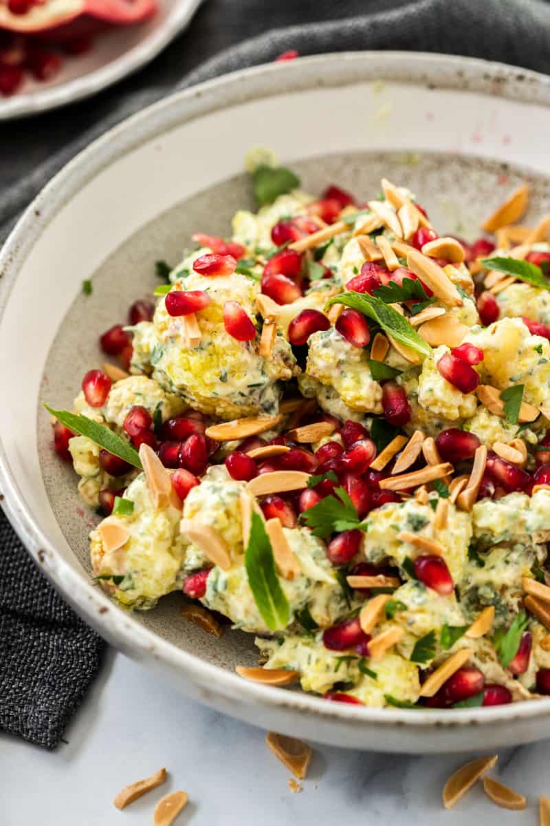 up close shot of bowl of cauliflower salad, sitting on a blue cloth. some slivered almonds are sprinkled at the front, and half a pomegranate is toward the back of the picture. 