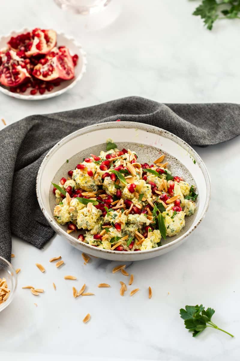 bowl of cauliflower salad, sitting on a blue cloth. some slivered almonds are sprinkled at the front, along with a small piece of parsley, and half a pomegranate is toward the back of the picture. photo taken on a white marble bench top.