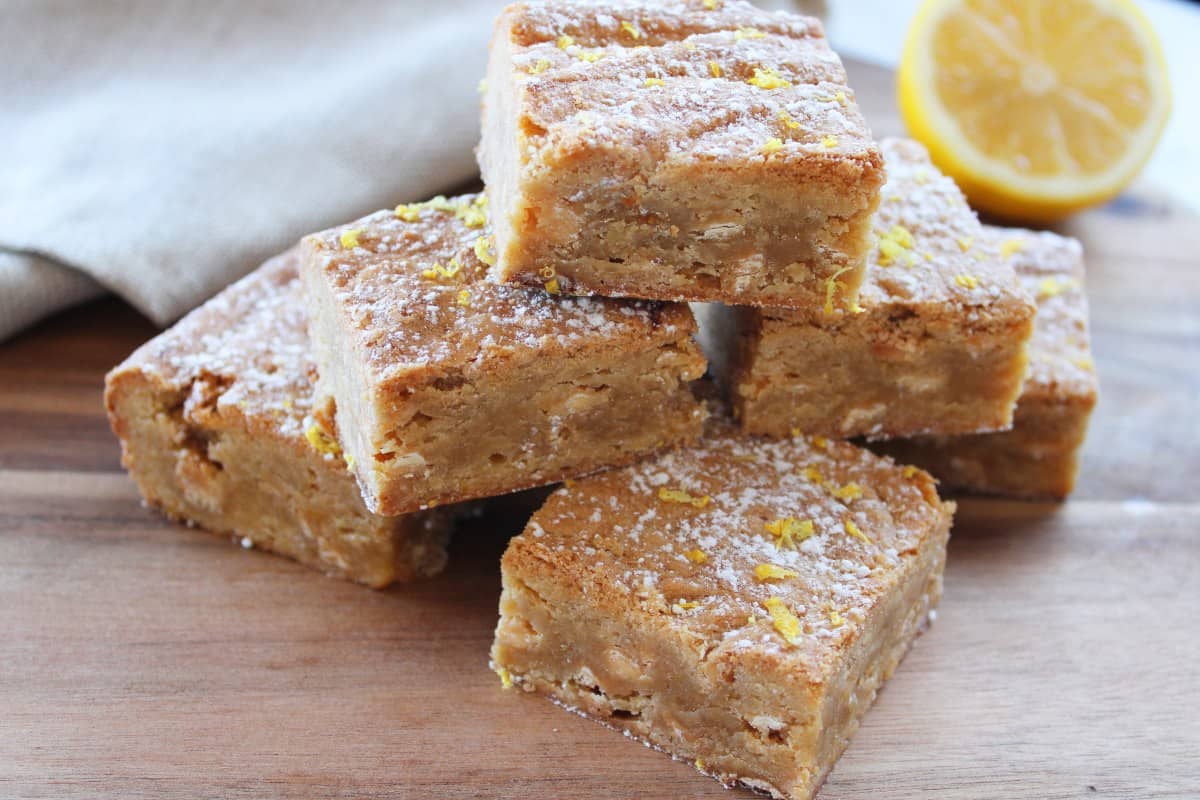 blondies stacked on round wooden board, with a beige napkin and half a cut lemon in the background.