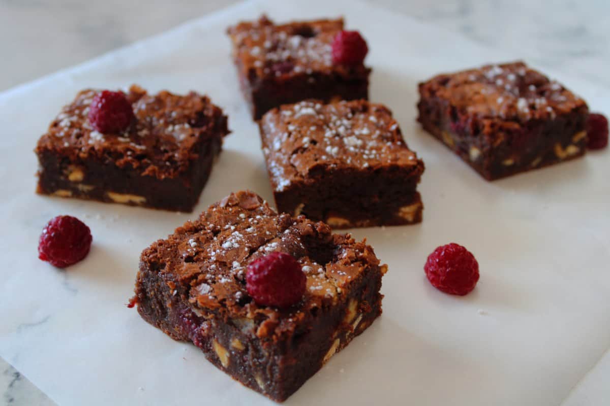 squares of brownies sitting on baking paper, dotted with some fresh raspberries.