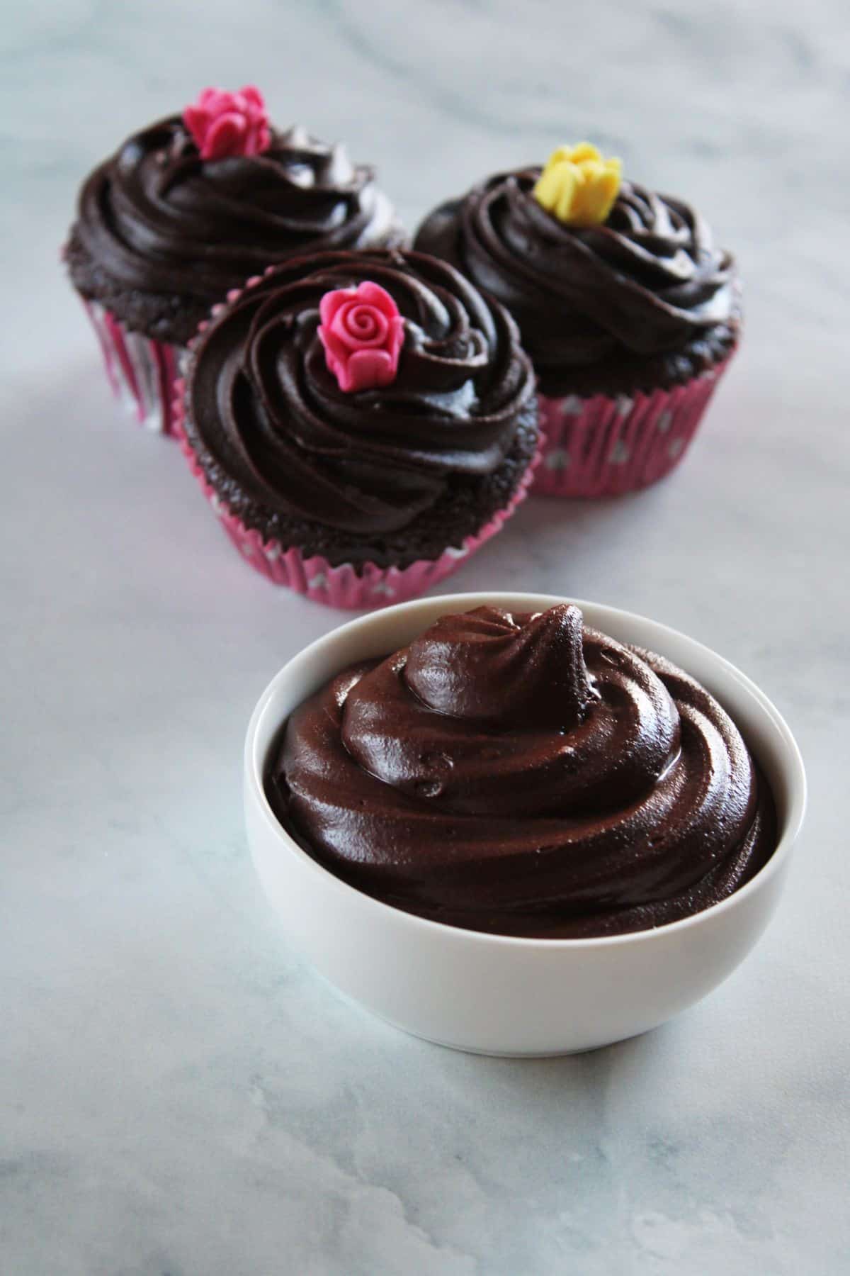 small white dish of chocolate frosting, with three cupcakes in the background, on a white marble benchtop.