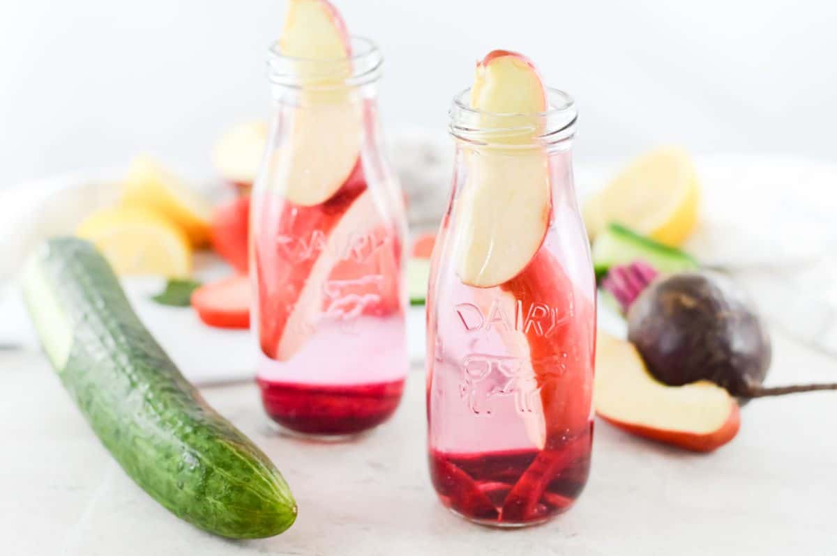 Two glass bottles on infused water (with beetroot and apple) on a board, surrounded by fruit and vegetables.