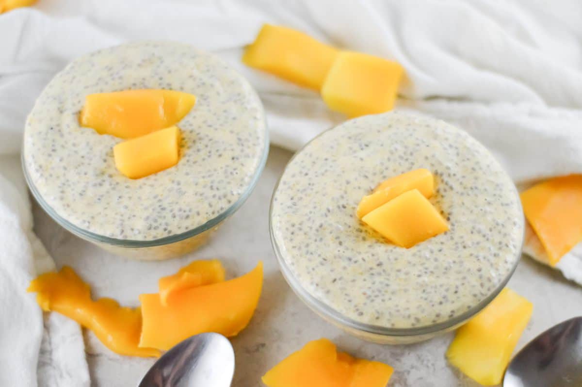 On a white bench, two bowls of chia pudding, garnished with mango sit. Mango pieces scattered around the bowls, and there are two silver spoons on the left. A white cloth in the background.