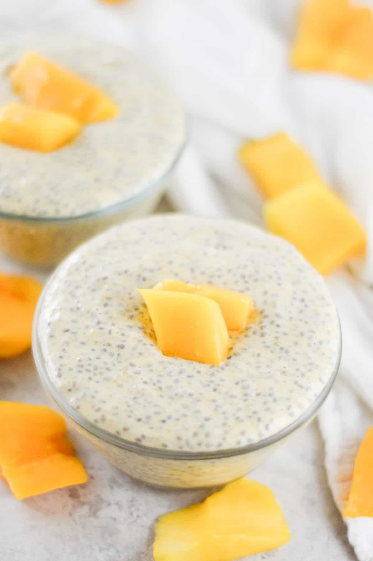 On a white bench, two bowls of chia pudding, garnished with mango sit. Mango pieces scattered around the bowls. A white cloth in the background.