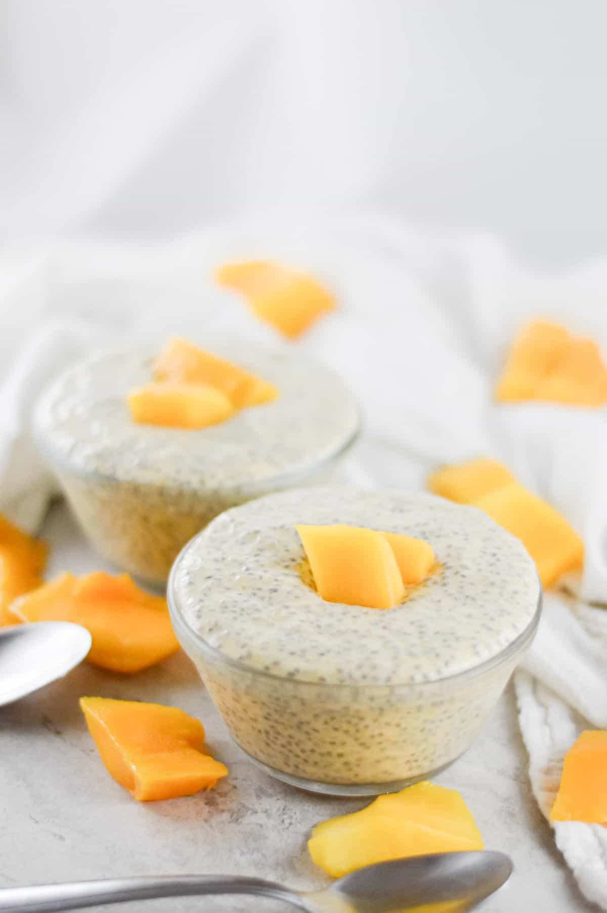 On a white bench, two bowls of chia pudding, garnished with mango sit. Mango pieces scattered around the bowls, and there are two silver spoon on the left. A white cloth in the background.