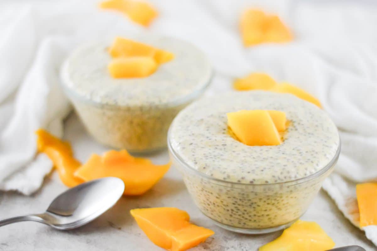 On a white bench, two bowls of chia pudding, garnished with mango sit. Mango pieces scattered around the bowls, and there is a silver spoon on the left. A white cloth in the background.