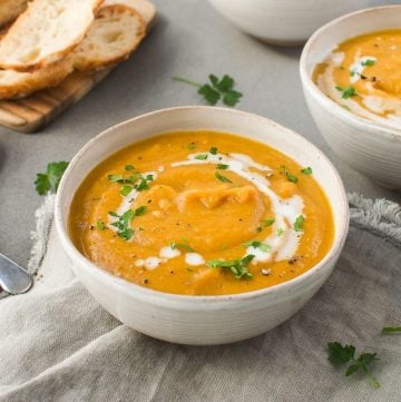 up close bowl of soup on a grey cloth, another bowl in the background to the right, some sliced bread to the left and two silver spoons on the side.